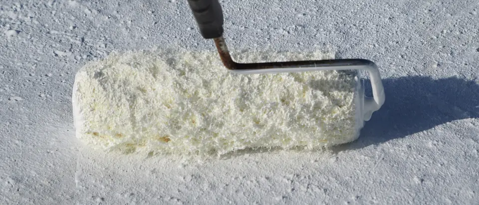 A paint roller on a pole spreading white coating over the rough surface of a flat roof