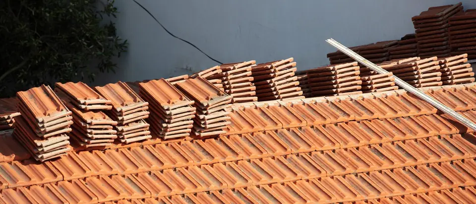 Stacks of clay roof tiles set out in a row along a tile roof, in front of a plain grey wall