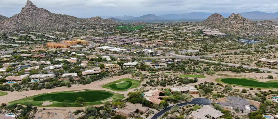 Flat-roofed and tile-roofed desert homes around golf greens in north Scottsdale, seen from the air, with a granite peak and boulder hills behind