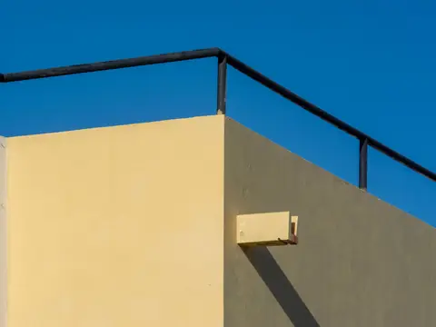A drain spout projecting through the stucco parapet wall of a flat-roofed building, casting a long shadow under a deep blue sky
