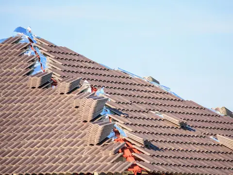 Stacks of concrete roof tiles lined up along the hip of a tile roof that is part way through being laid, under a pale blue sky