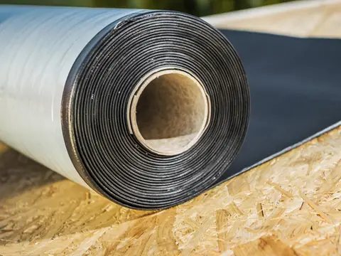 A roll of black roofing membrane partly unrolled across a bare wood roof deck